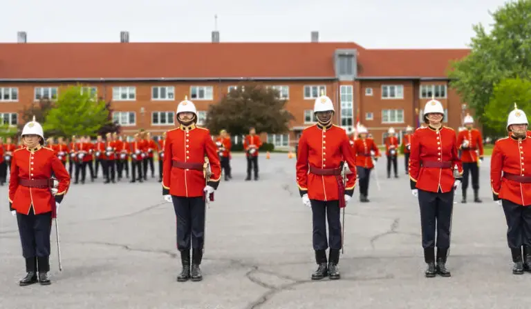 Un groupe de personnes v&ecirc;tues d'uniformes militaires rouges et de casques blancs se tient en formation sur une esplanade pav&eacute;e, tandis que d'autres, portant une tenue similaire, se tiennent &agrave; l'arri&egrave;re-plan pr&egrave;s d'un b&acirc;timent au toit rouge.