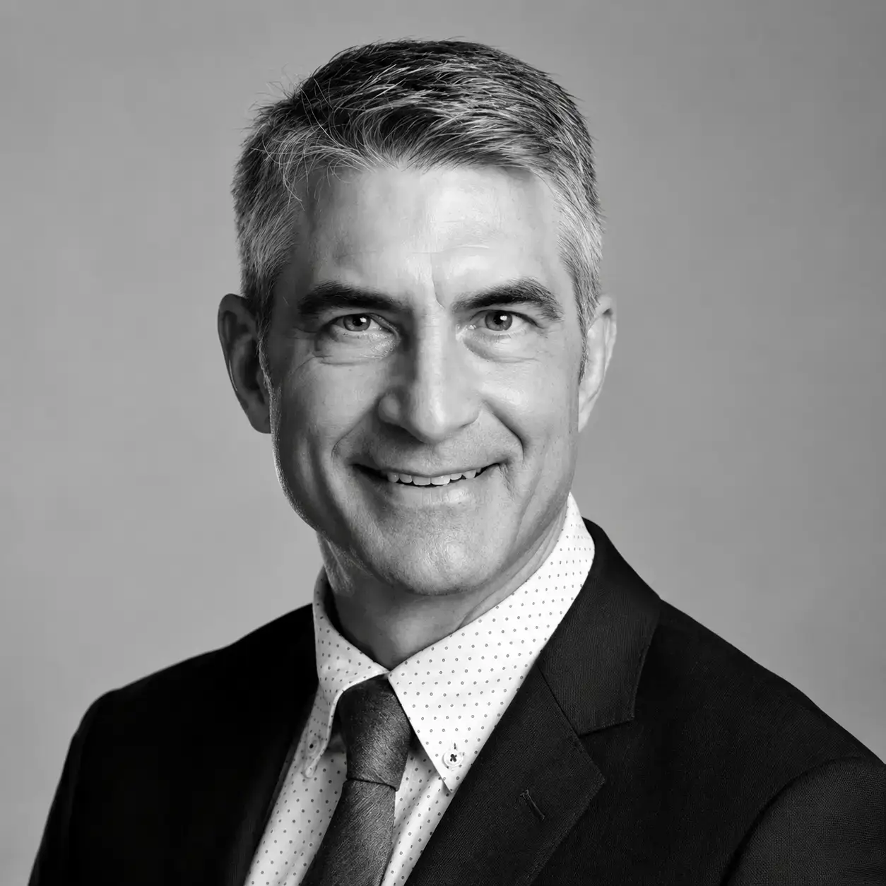 Portrait en noir et blanc d'un homme d'âge mûr aux cheveux courts et gris, vêtu d'un costume, d'une chemise blanche à petits motifs et d'une cravate, souriant à l'appareil photo devant un fond uni.