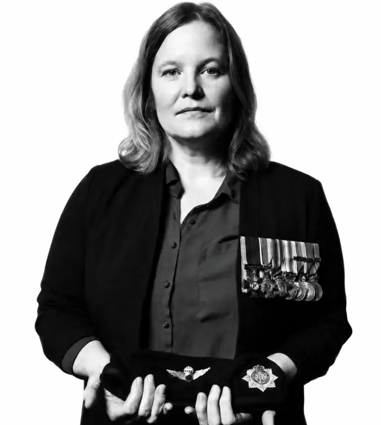 A woman with medals on her jacket holds a beret and a military badge in her hands, facing the camera with a serious expression against a plain white background.