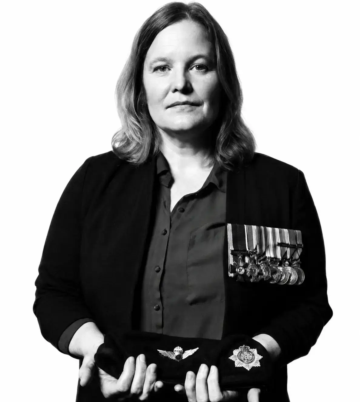 A woman with medals on her jacket holds a beret and a military badge in her hands, facing the camera with a serious expression against a plain white background.