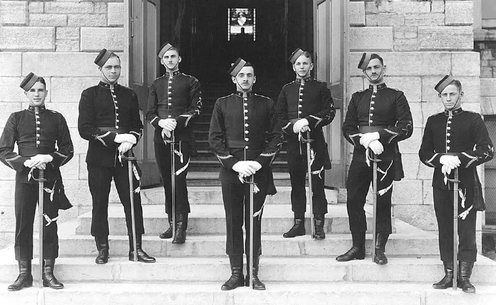 Seven men in military uniforms and hats stand in a formal pose on stone steps outside a building, each holding a sword and wearing white gloves. The photo is black and white and appears historical.