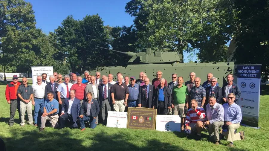 A large group of men pose in front of a military tank monument outdoors on grass, with commemorative plaques and project signs nearby under sunny skies and trees.