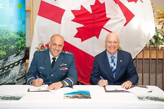 Deux hommes en costume sont assis à une table, en train de signer des documents, avec un grand drapeau canadien en arrière-plan. Tous deux sourient ; l'un porte un uniforme militaire, l'autre un costume et une cravate.