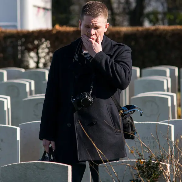 A man with a camera around his neck stands among gravestones, covering his mouth with his hand, appearing emotional or thoughtful.