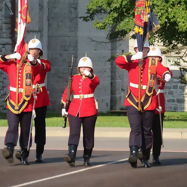 Four uniformed guards in red jackets and white helmets march in formation, carrying flags. They are on a paved road, with a stone building and trees visible in the background.