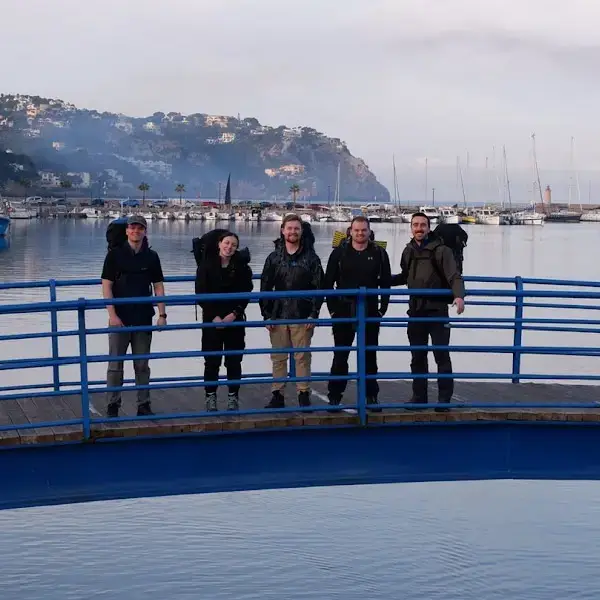 Cinq personnes, munies de sacs à dos, se tiennent sur un pont bleu incurvé enjambant un cours d'eau, avec en arrière-plan des bateaux et une rive vallonnée, sous un ciel nuageux.