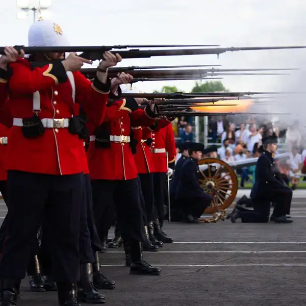 Des soldats en uniforme rouge tirent à l'arme à feu en formant un salut synchronisé, tandis qu'à l'arrière-plan, des hommes en uniforme sombre actionnent un canon, d'où s'échappe de la fumée. Des spectateurs assistent à la scène depuis les tribunes.