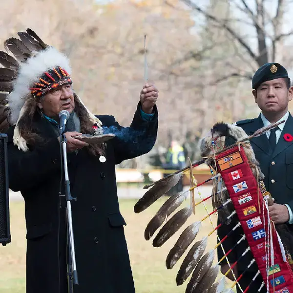 An Indigenous elder in a feathered headdress holds a feather and smudge bowl while speaking at a microphone. A uniformed soldier stands beside him, holding an eagle staff adorned with flags.