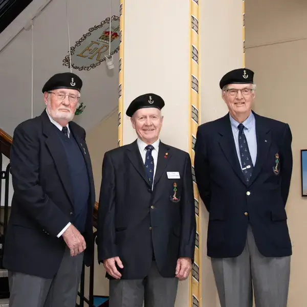 Three elderly men in matching navy blazers, blue shirts, and black berets with anchor insignias stand together indoors, smiling at the camera near a staircase.
