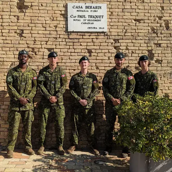 Cinq soldats canadiens en uniforme se tiennent devant un mur de briques et une plaque commémorative. La lumière du soleil projette des ombres. On aperçoit du feuillage vert dans le coin inférieur droit.