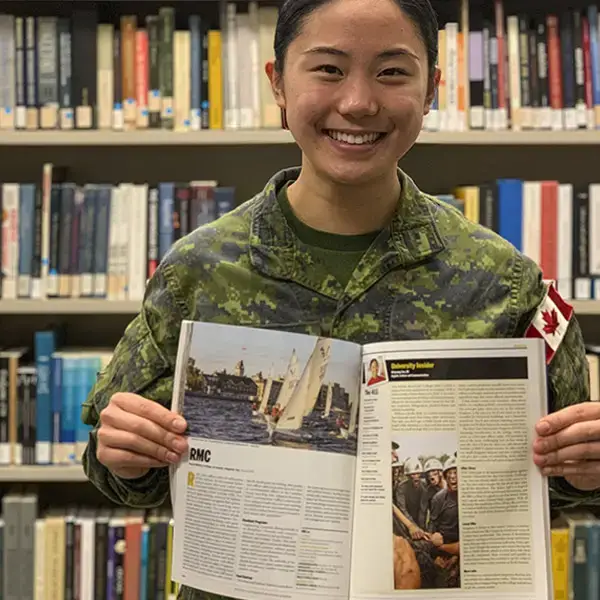 A person in military uniform smiles while holding open a magazine in a library. The magazine features an article with photos, and bookshelves filled with books are visible in the background.