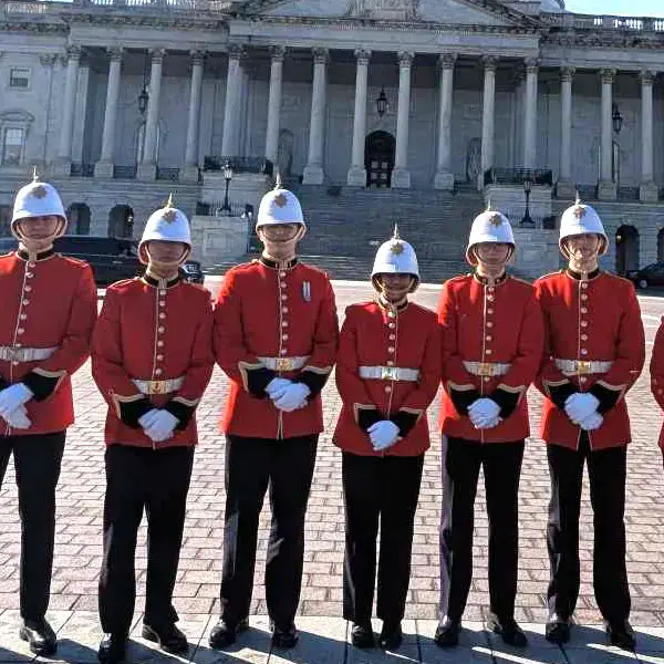 Seven people in red British-style ceremonial uniforms and white helmets stand in a line outside a large, columned building on a sunny day.