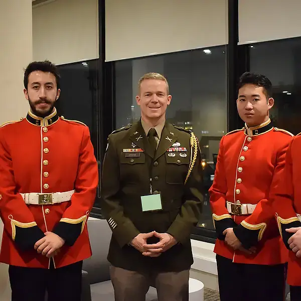 Three men in formal military uniforms stand indoors, two in red jackets with white belts and one in an olive green uniform, posing and smiling for a photo in front of large windows at night.