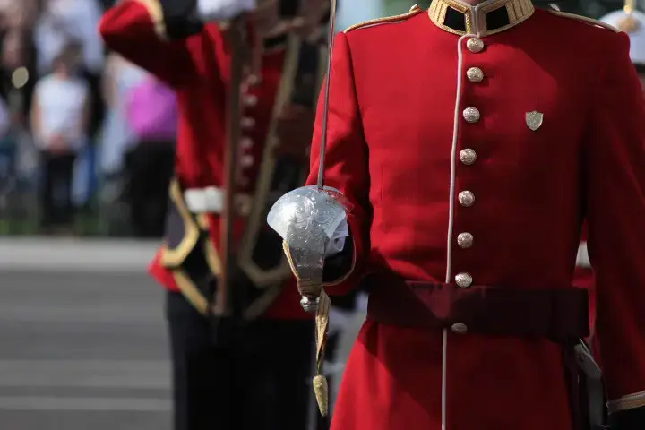 Gros plan sur un garde en uniforme, vêtu d'un manteau rouge et tenant une épée, avec un autre garde flou à l'arrière-plan, lors d'une cérémonie.