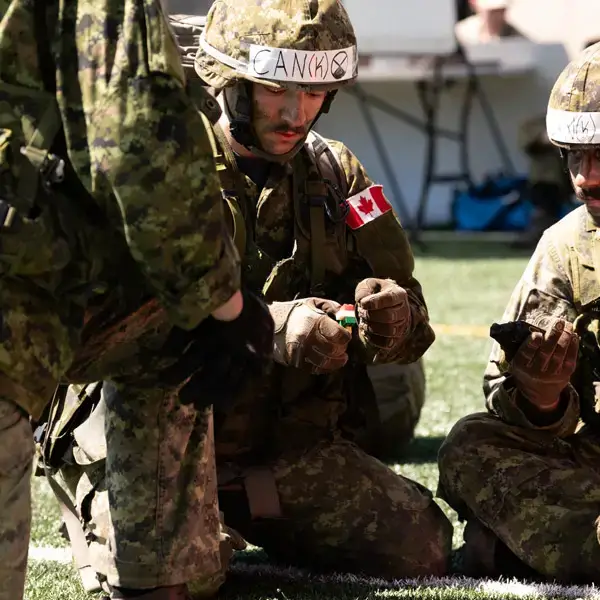 Trois soldats en tenue de camouflage, portant sur leurs casques des étiquettes « CAN(8) » et des écussons représentant le drapeau canadien, sont agenouillés sur l'herbe et manipulent des objets lors d'un entraînement ou d'un exercice sur le terrain.