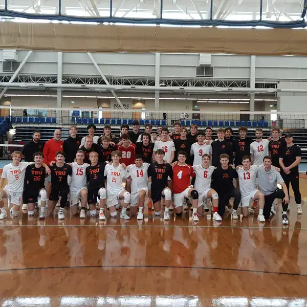 Photo de groupe d'une équipe masculine de volley-ball sur un terrain couvert ; les joueurs portent des tenues TRU blanches ou noires, des maillots numérotés, certains sont à genoux et d'autres debout, avec les entraîneurs à leurs côtés.