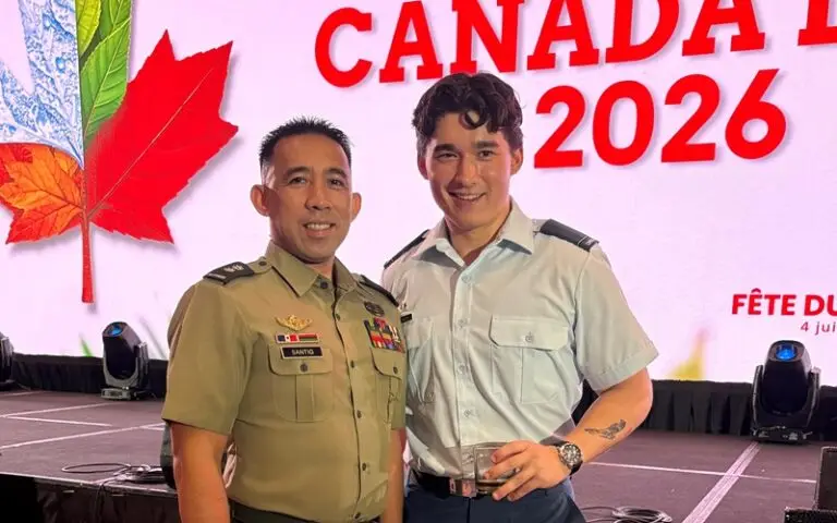 Two men in military uniforms smile on stage. A large screen behind them displays a red maple leaf and the words “CANADA DAY 2026.” Stage lights and technical equipment are visible in the background.