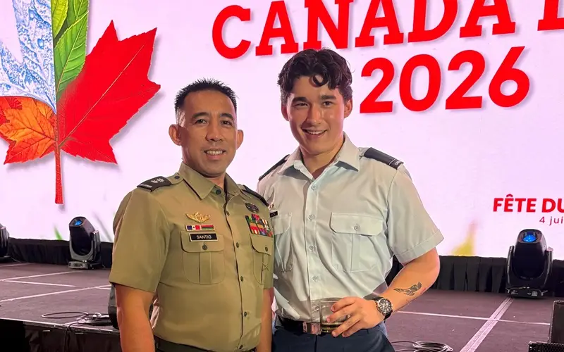 Two men in military uniforms smile on stage. A large screen behind them displays a red maple leaf and the words “CANADA DAY 2026.” Stage lights and technical equipment are visible in the background.