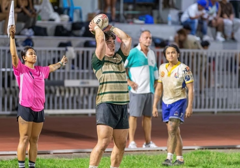 A rugby player prepares for a line-out throw, flanked by an official in pink holding a flag and two other players. Spectators sit in the stands behind a metal fence.