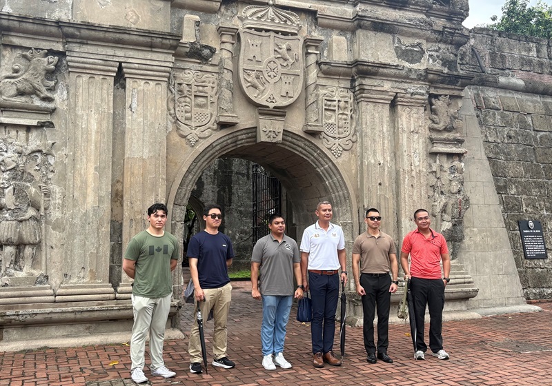 Six men stand in front of the stone entrance arch of Fort Santiago in Manila, featuring detailed carvings and historical emblems above the gate. The group poses for a photo on brick pavement.