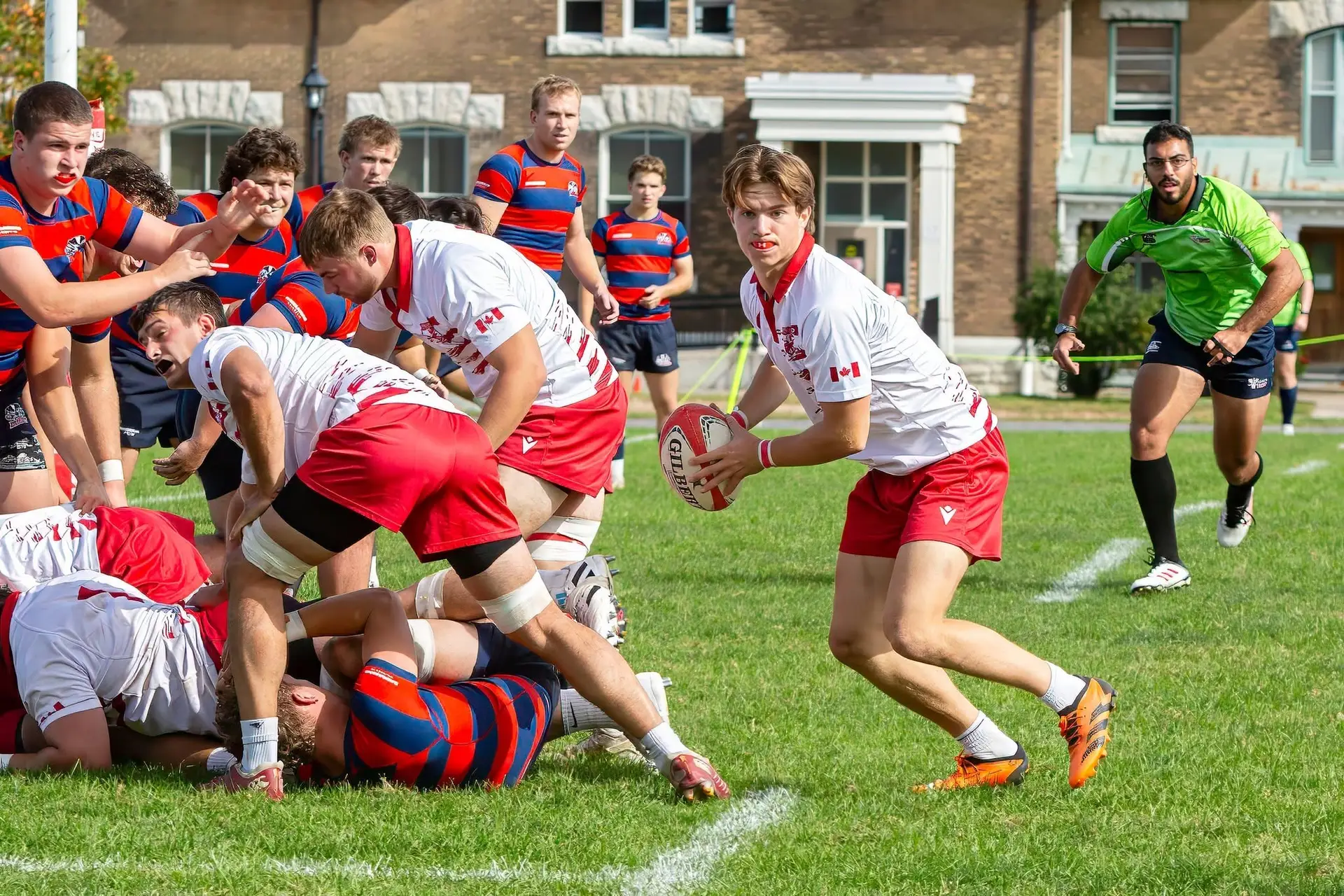 A rugby player in a white and red kit holds the ball, ready to pass, whilst teammates and opponents in blue and red stripes engage behind him on a grassy pitch.