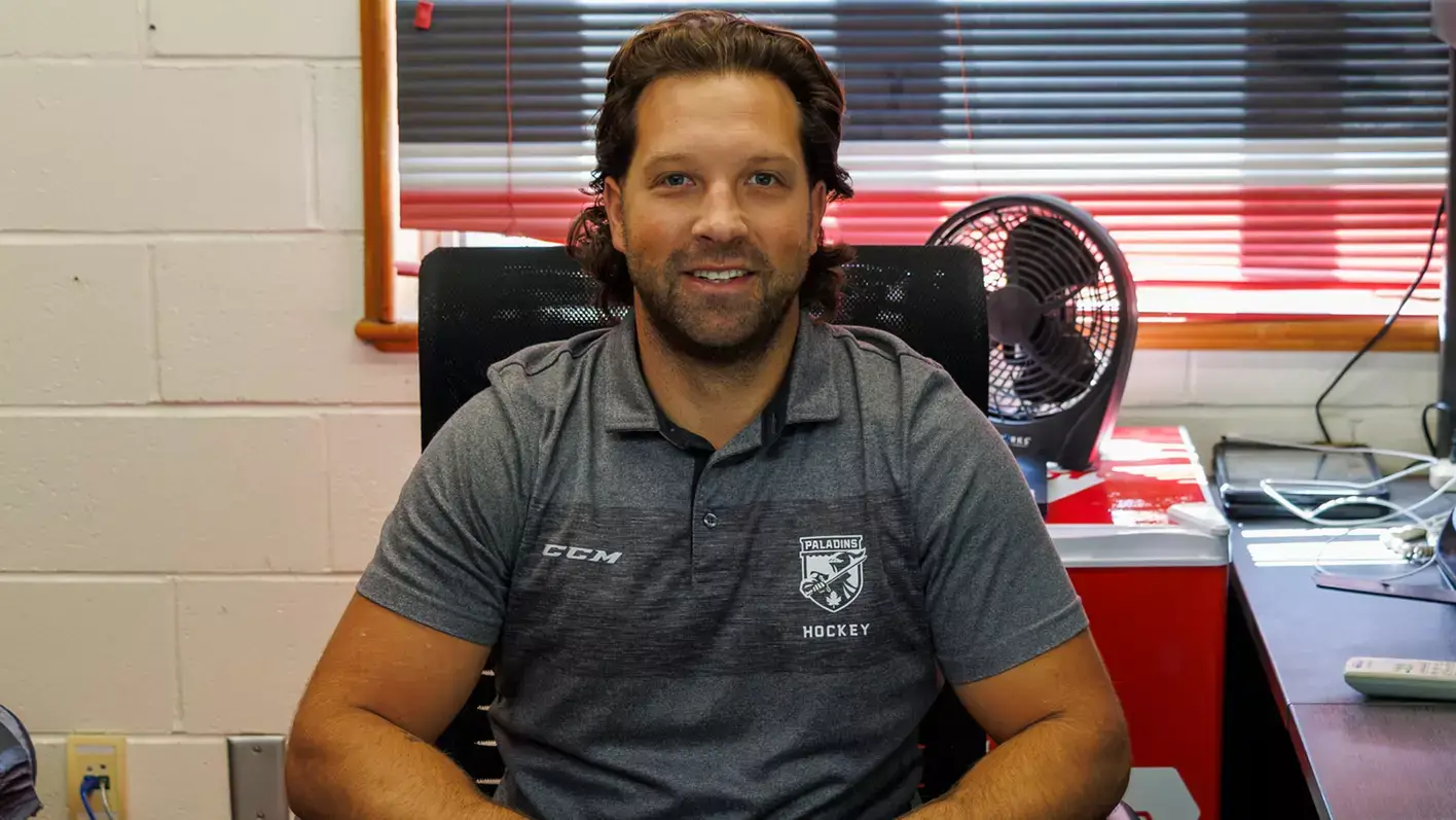 A man with long brown hair and a beard sits at a desk, smiling. He wears a grey hockey polo shirt. Behind him are a fan, office supplies, and a window with blinds.