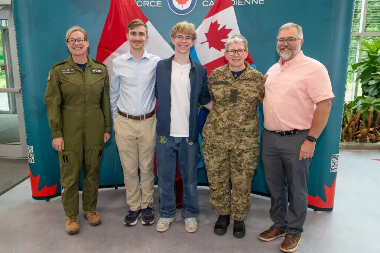 Five people stand smiling in front of Canadian flags and a blue backdrop. Two wear military uniforms, two are in casual clothing, and one is in business casual. They stand close together in a bright indoor space.