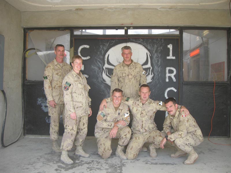 Six Canadian soldiers in camouflage uniforms pose for a photo in front of a painted wall featuring a skull emblem and the letters “C” and “R”.