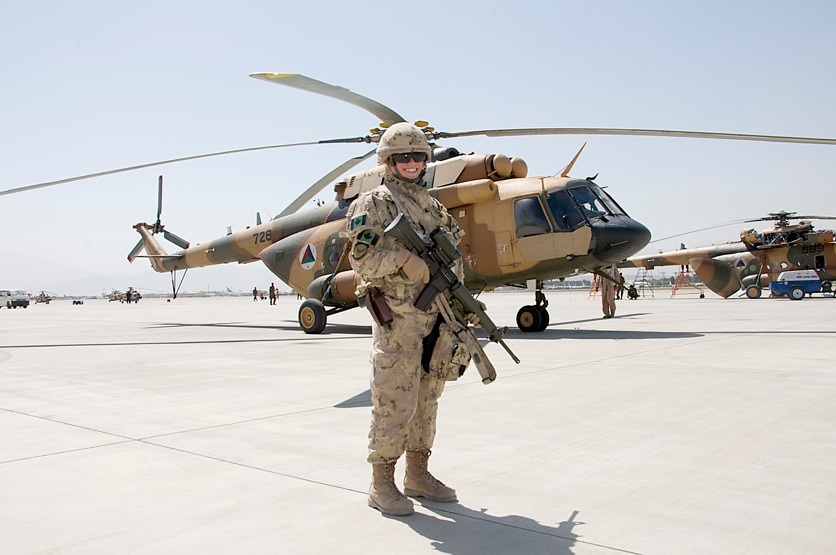 A smiling Canadian soldier in desert camouflage uniform stands on an airfield in front of a parked military helicopter.