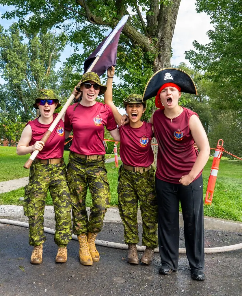 Four people in matching maroon shirts and camouflage trousers posing and cheering outdoors, with one person wearing a pirate hat and another holding a flag.