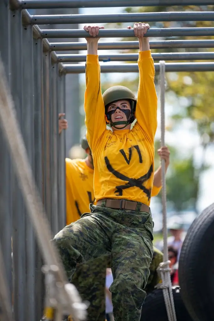 A soldier in a yellow long-sleeved shirt and camouflage trousers navigates a set of monkey bars on an assault course.