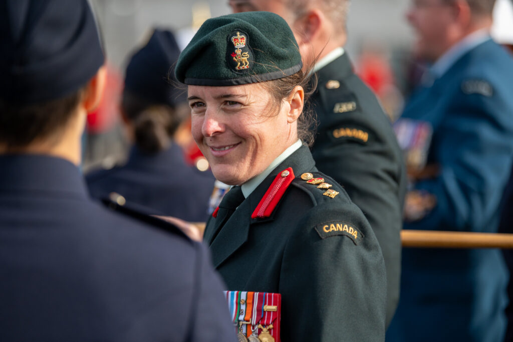 A Canadian military officer in a green beret and uniform smiles whilst speaking to another person in uniform.