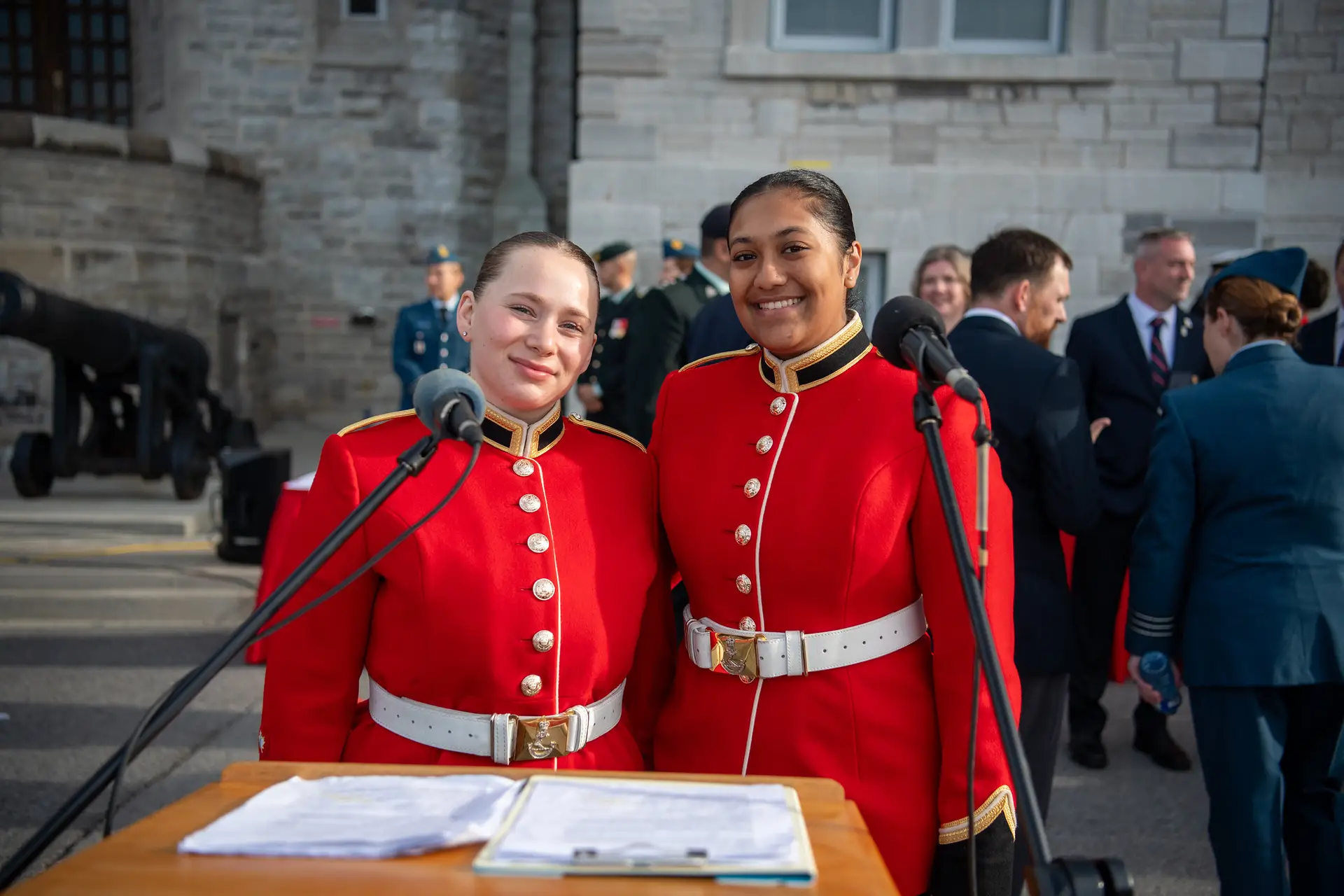 Two Canadian Armed Forces members in scarlet ceremonial uniforms stand behind a lectern with microphones, smiling at the camera.