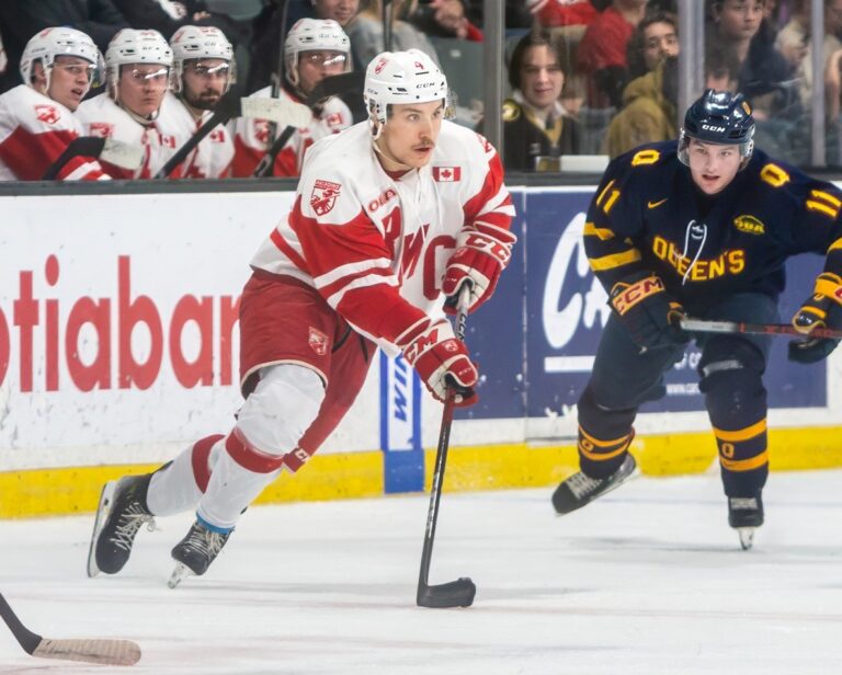 A hockey player in a white jumper with red trim skating with the puck, pursued by an opponent in a dark blue and gold jumper.
