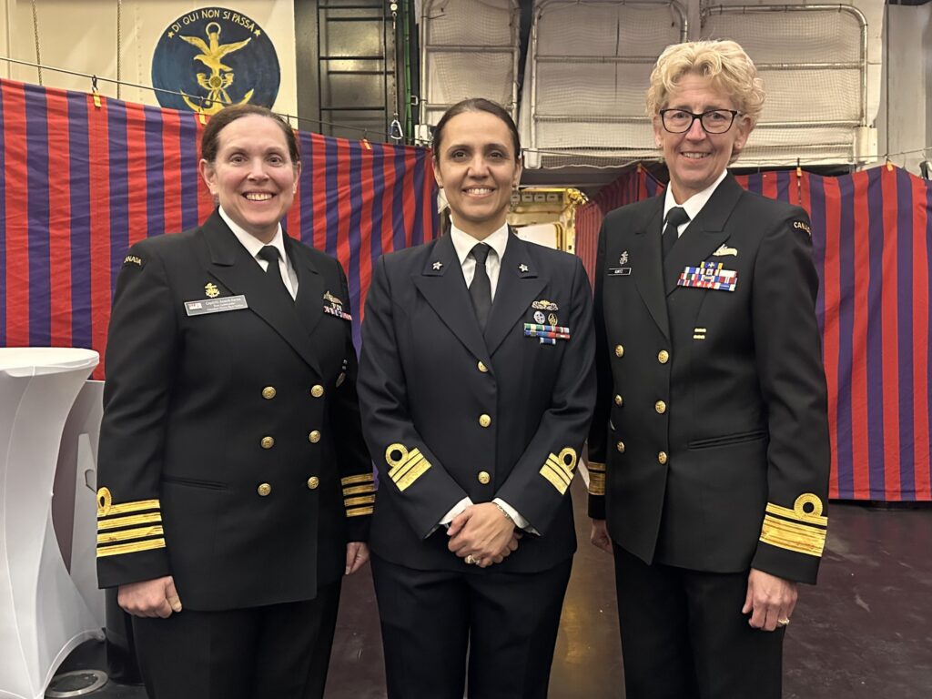 Three Canadian Navy officers in formal dark uniforms standing side-by-side in front of a blue and red striped curtain.