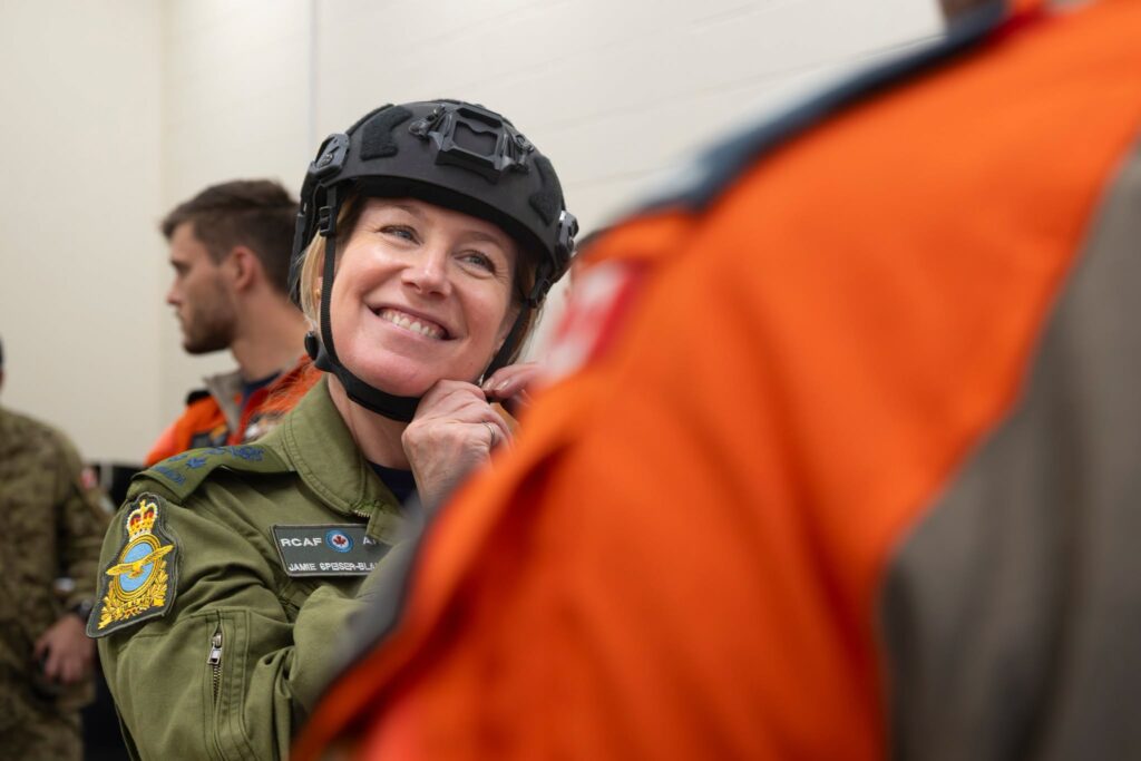 A smiling Royal Canadian Air Force officer in flying gear adjusts her helmet, with blurred figures of other personnel in the foreground and background.