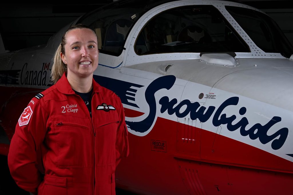 Pilot Caitie Clapp wearing a red flight suit stands in front of a Snowbirds aeroplane.