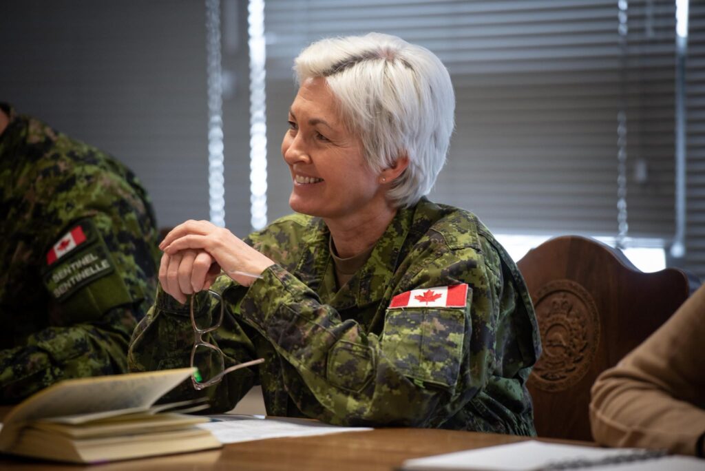 A Canadian Armed Forces officer with short white hair smiles whilst sitting at a table during a meeting.