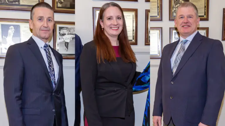 Three people in business attire stand indoors against a wall decorated with framed photos and documents, posing separately for formal portraits.
