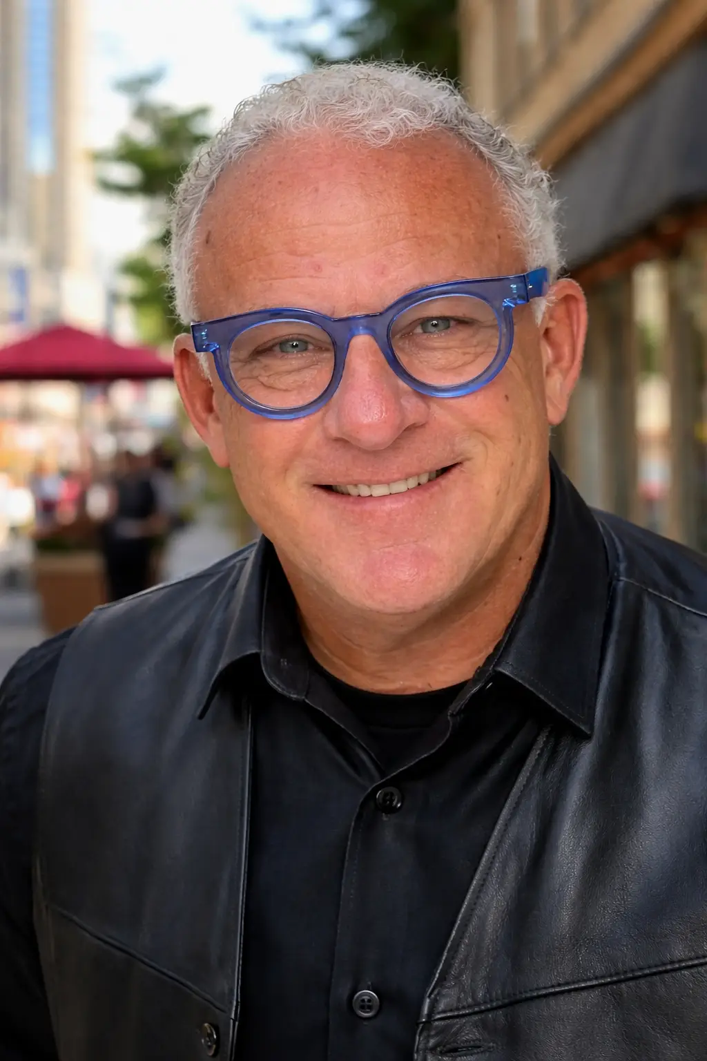 Smiling man with short curly grey hair and blue glasses wears a black leather jacket and shirt, standing outdoors on a city street with blurred buildings and umbrellas in the background.