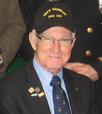 An older man wearing a suit, tie with anchor motifs, and a black baseball cap that reads “HMCS GATINEAU DDE 236”.