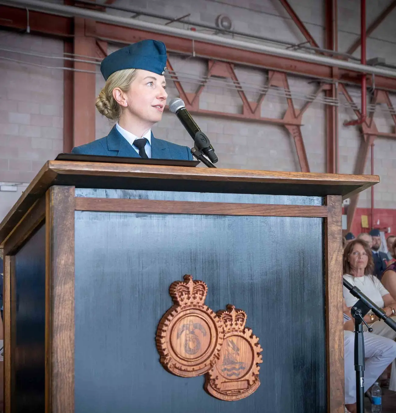 A female officer in a blue military uniform speaks into a microphone at a wooden lectern decorated with two carved crests.