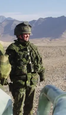 A soldier in camouflage uniform and helmet stands in a desert landscape with mountains in the background.
