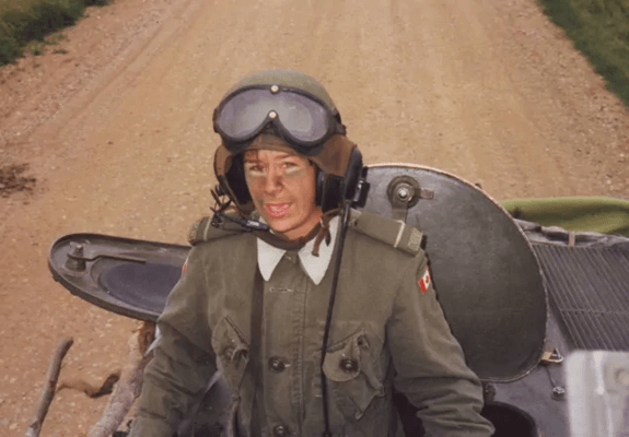 A Canadian soldier in uniform with camouflage paint and goggles sitting in the hatch of a military vehicle on a dirt track.