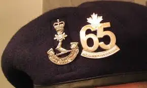 A dark blue beret featuring two metal military badges, one depicting a crown and crest, and the other displaying the number 65 beneath a maple leaf.