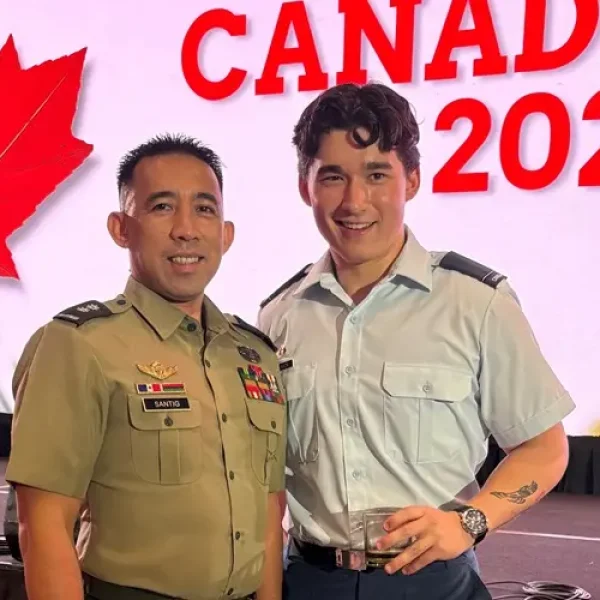 Two men in military uniforms smile on stage. A large screen behind them displays a red maple leaf and the words &ldquo;CANADA DAY 2026.&rdquo; Stage lights and technical equipment are visible in the background.