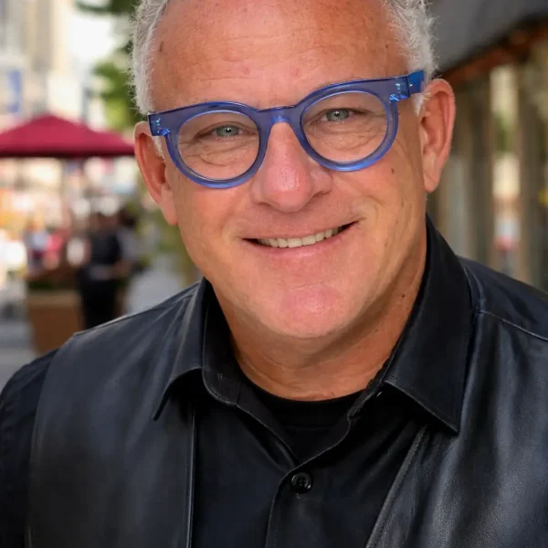 Smiling man with short curly grey hair and blue glasses wears a black leather jacket and shirt, standing outdoors on a city street with blurred buildings and umbrellas in the background.
