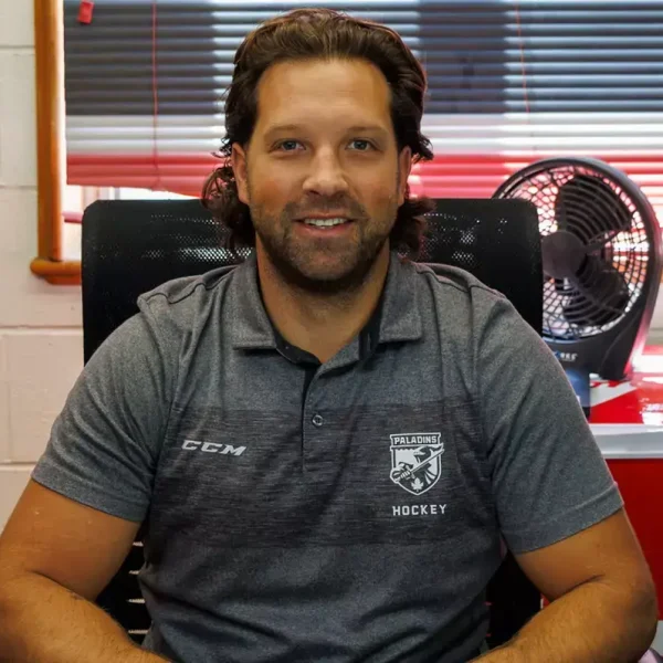 A man with long brown hair and a beard sits at a desk, smiling. He wears a grey hockey polo shirt. Behind him are a fan, office supplies, and a window with blinds.