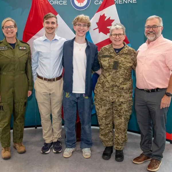 Five people stand smiling in front of Canadian flags and a blue backdrop. Two wear military uniforms, two are in casual clothing, and one is in business casual. They stand close together in a bright indoor space.