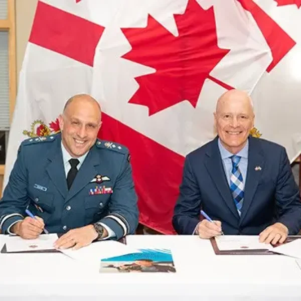 Two men in suits sit at a table signing documents, smiling toward the camera. A large Canadian flag hangs behind them. Official documents and pens are on the table.